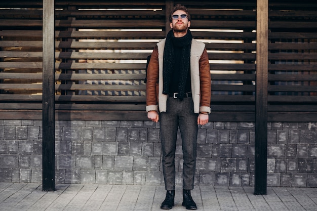 A stylish man wearing a brown jacket, scarf, and gray trousers stands outdoors near a wooden structure.