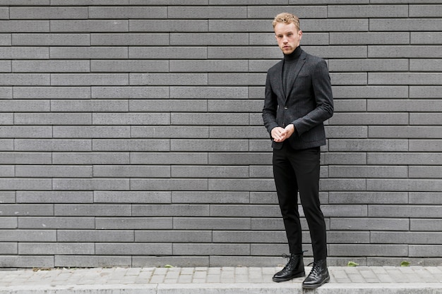 A man in a sleek black suit and turtleneck stands confidently against a modern gray brick wall.
