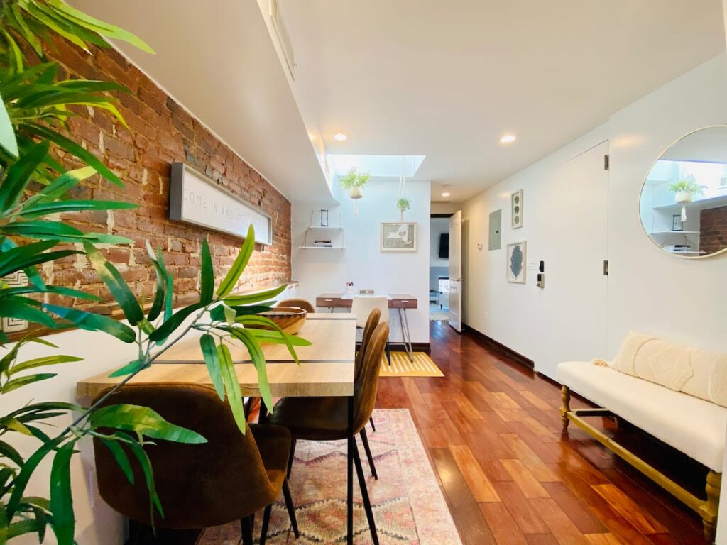 A modern, cozy living space with a brick wall, wooden floor, dining area, and white sofa, featuring plants and warm lighting.