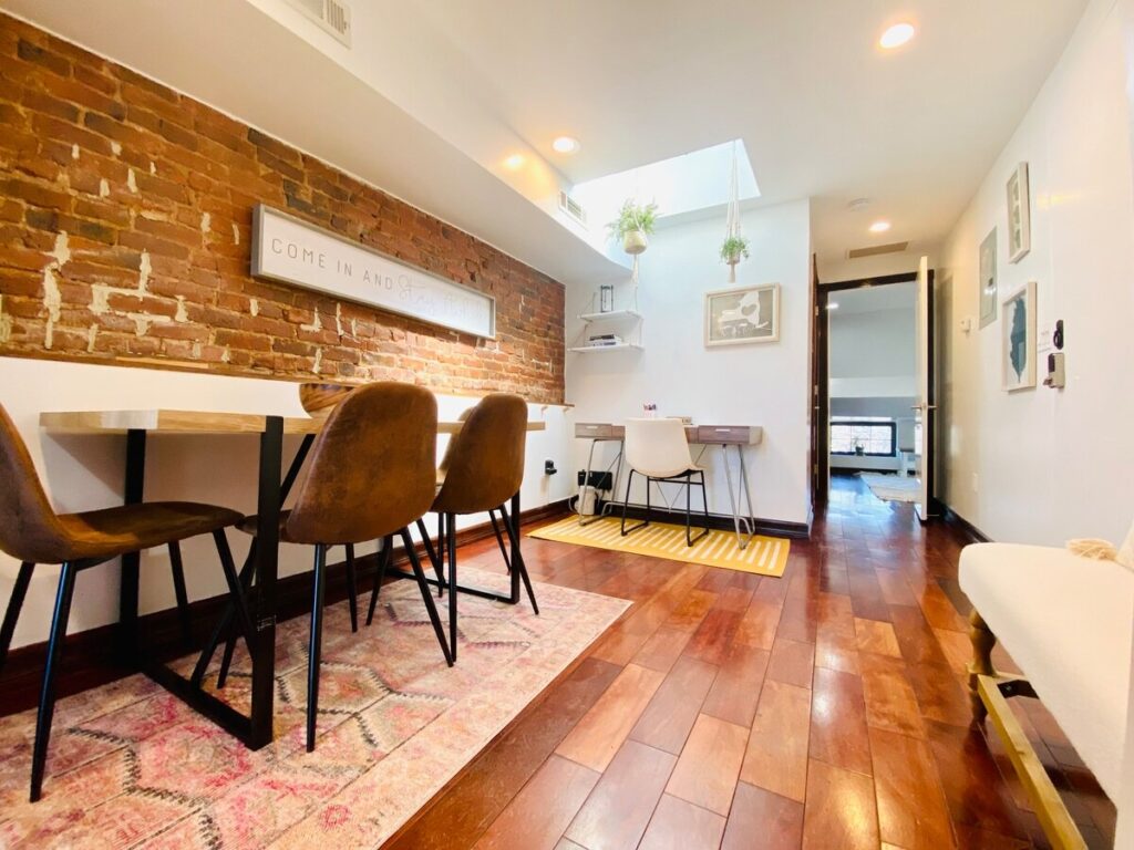 A bright, modern living space with a brick wall, wooden floor, dining area, and white sofa, accented by plants and warm lighting.