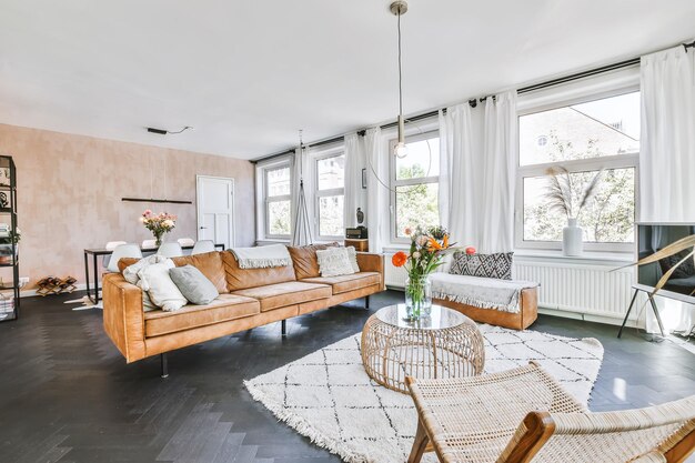 Bright and airy living room featuring a tan leather sofa, woven chairs, a round wicker coffee table, and large windows with white curtains letting in natural light.