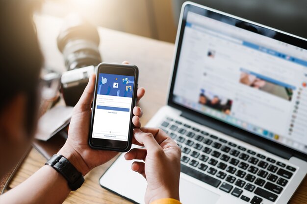 A person holding a smartphone displaying the Facebook login screen, with a laptop open to the Facebook website in the background. A camera lens and leather wallet are on the wooden desk beside them.