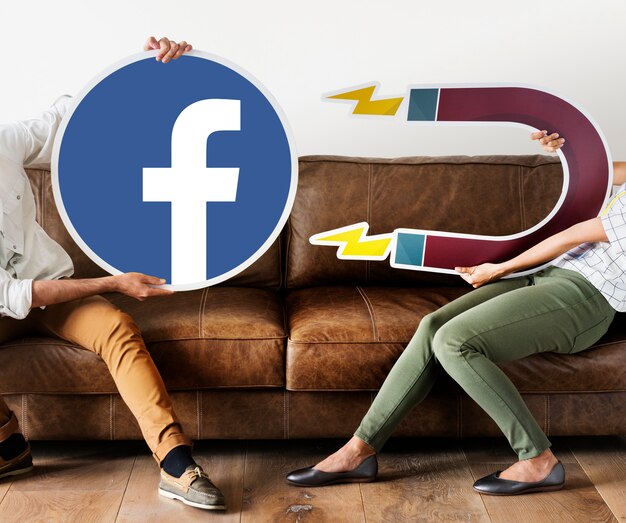 Two people sitting on a brown leather couch, one holding a large Facebook logo cutout and the other holding a giant magnet illustration with lightning bolts, symbolizing attracting followers or engagement on Facebook.