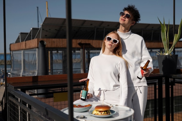 A stylish couple in matching white outfits enjoying drinks on a rooftop restaurant with food and modern decor in the background.