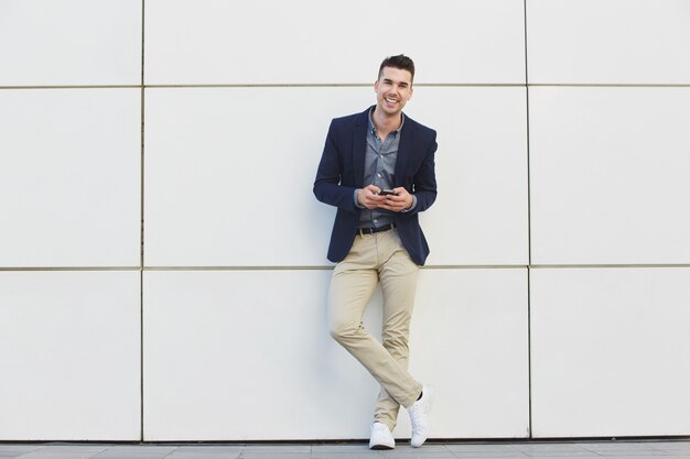 Smiling man in a navy blazer, gray shirt, and beige pants leaning against a white wall while holding a smartphone.
