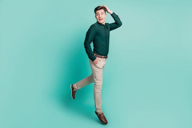 Smiling man in a dark green shirt and beige pants posing playfully on a turquoise background with one leg lifted.