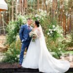 Bride and groom beneath a floral arch in a forest wedding setting, holding a bouquet, surrounded by candles and soft light.