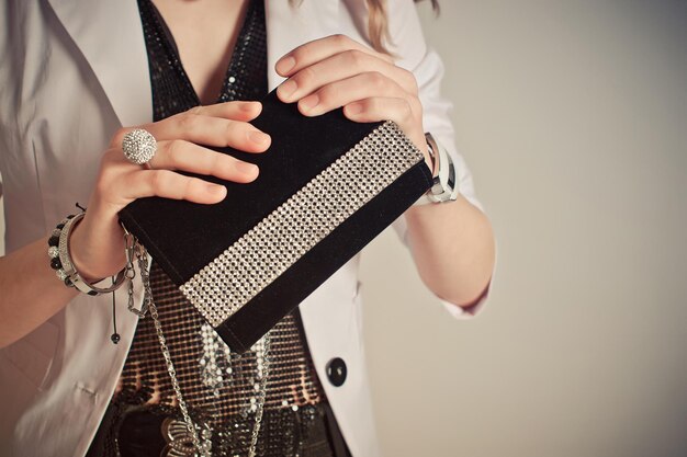 A woman holding a black clutch purse with a sparkling rhinestone stripe, wearing multiple rings and bracelets.