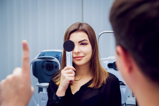 Young woman taking a vision test in an eye clinic, holding an occluder while the eye specialist checks focus and clarity.