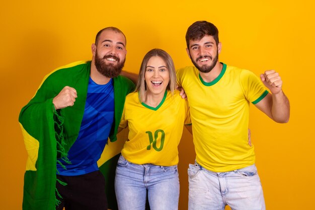 Three Brazil fans in yellow jerseys smiling and cheering against a bright yellow background.